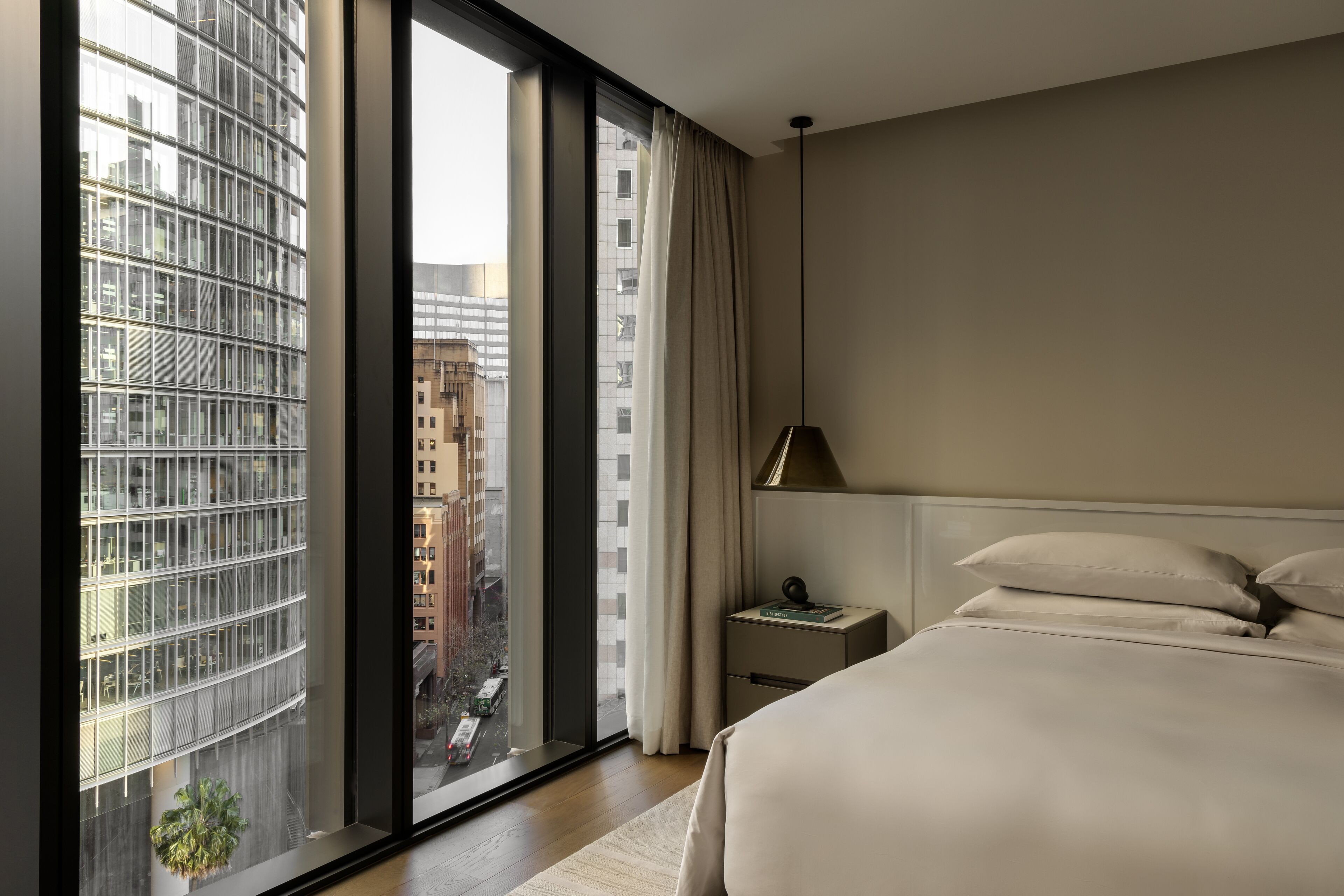 A modern hotel room with a large window, a bed, a nightstand, and a lamp.