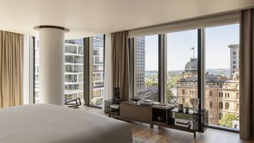 Una habitación de hotel moderna con una cama grande, un escritorio con vistas y un paisaje urbano visible a través de ventanas amplias.