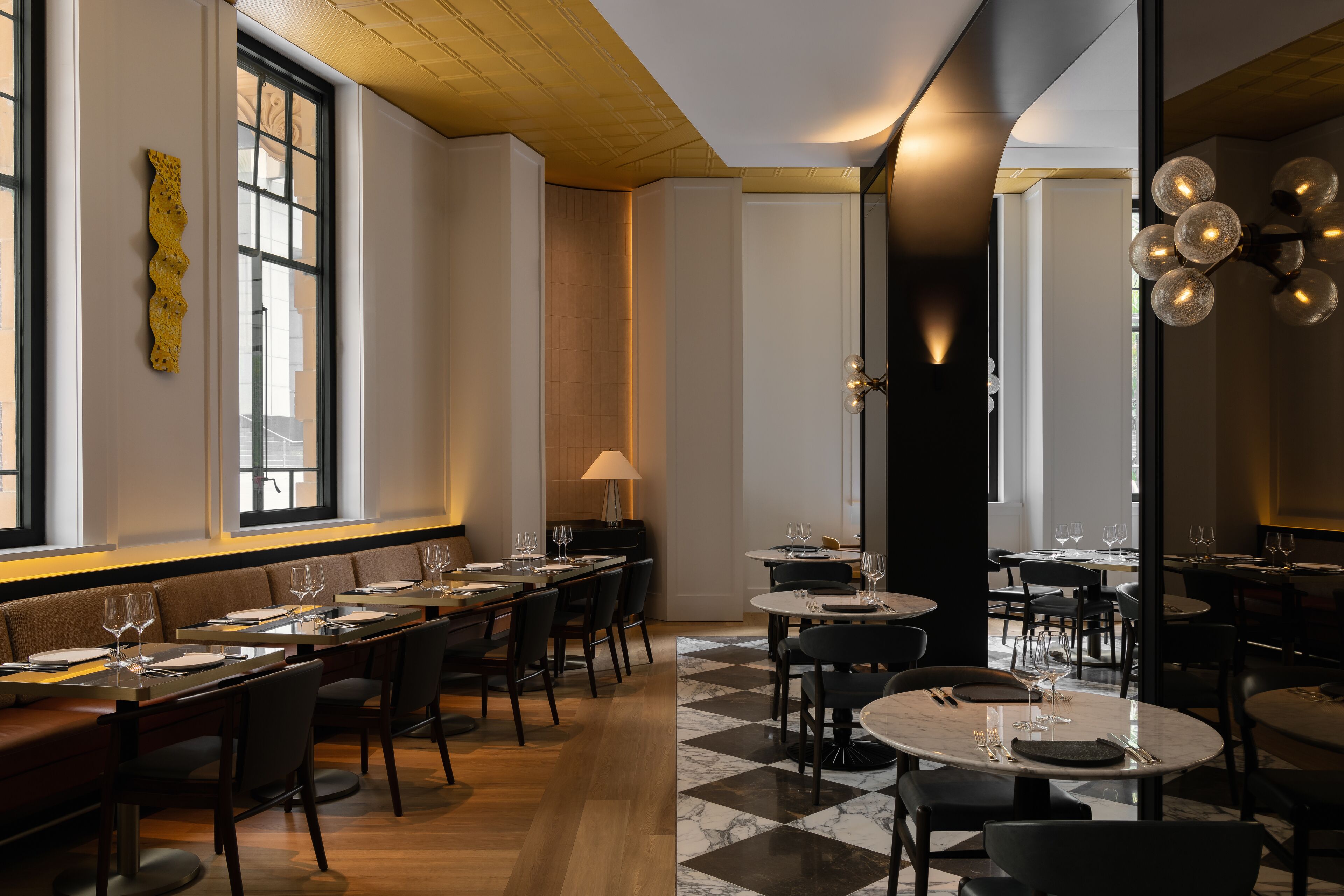 Un restaurant avec un sol à carreaux, de longues tables et des chaises. Il y a des fenêtres avec des stores, un luminaire fixé au mur et un élément décoratif mural.