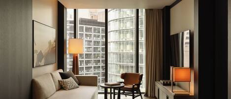 Une chambre d’hôtel moderne équipée d’un canapé, d’une petite table, d’une chaise, d’un téléviseur et d’une grande fenêtre donnant sur la ville.