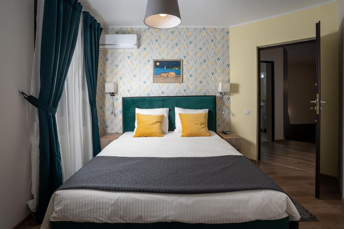 Ein ordentlich bezogenes Bett mit einem grünen Kopfteil, zwei Kissen und einer grauen Decke. Darüber an der Wand hängt ein Gemälde, und zur Rechten befindet sich eine Tür.