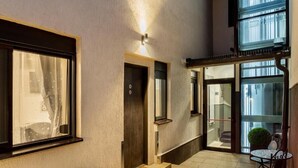 Un moderno vestíbulo de hotel con una escalera que conduce a un piso superior, un pequeño área de descanso con una mesa y sillas, y vistas de la ciudad a través de amplias ventanas.