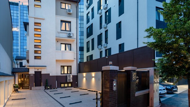 Un edificio moderno de varios pisos, con entrada protegida y un estacionamiento con varios coches.