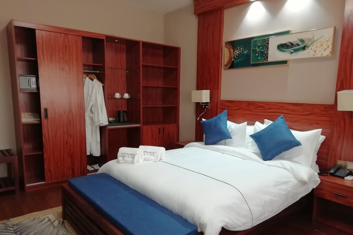 A hotel room with a bed, a wooden wardrobe, a bench, a bedside table with a phone, and a wall-mounted artwork.