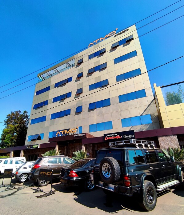 Un hotel de varios pisos con un cartel de cajero automático destacado, un estacionamiento con varios coches y un área de estar al aire libre visible.