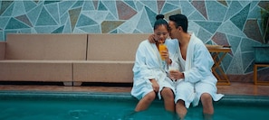 Dos personas sentadas junto a una piscina, vestidas con batas blancas, con una pared de mosaicos y un banco de madera al fondo.