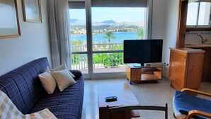 Een woonkamer met een bank, een tv op een dressoir, een klein tafeltje en een balkon met uitzicht op zee en bergen.
