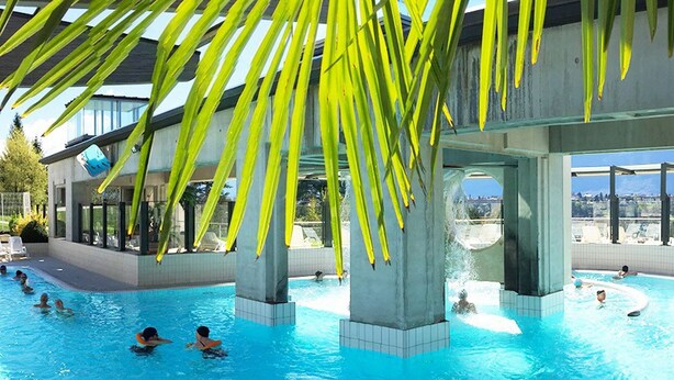 Una piscina con agua azul cristalina, rodeada de pilares de concreto y paredes de vidrio, con vistas al paisaje urbano al fondo.