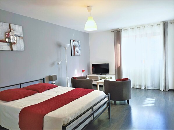 A modern hotel room with a large bed, a desk, a chair, a flat-screen TV, and a large window with curtains.