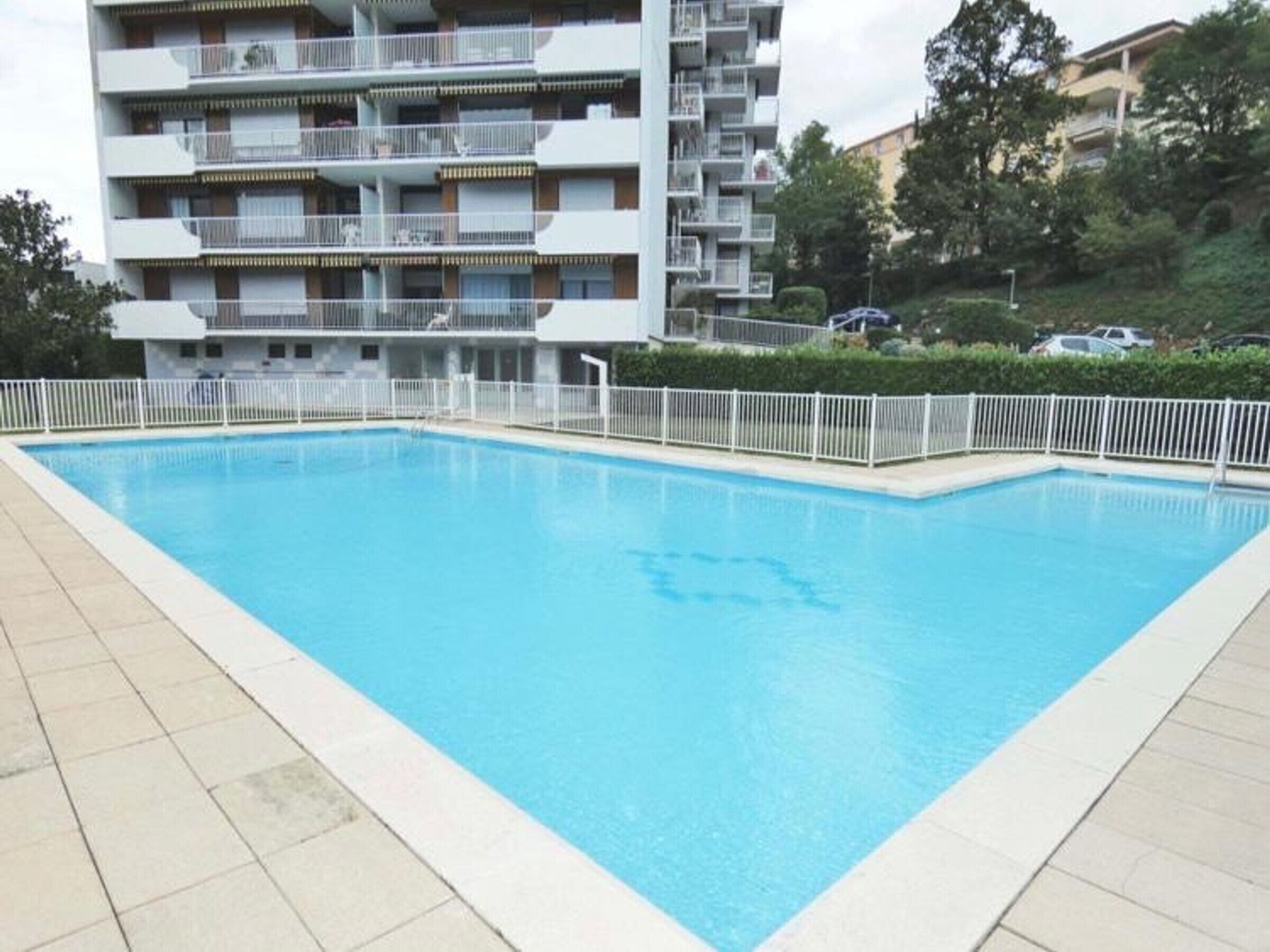 Una piscina rectangular amb una tanca blanca al seu voltant, amb un edifici residencial de diverses plantes i balcons com a fons.