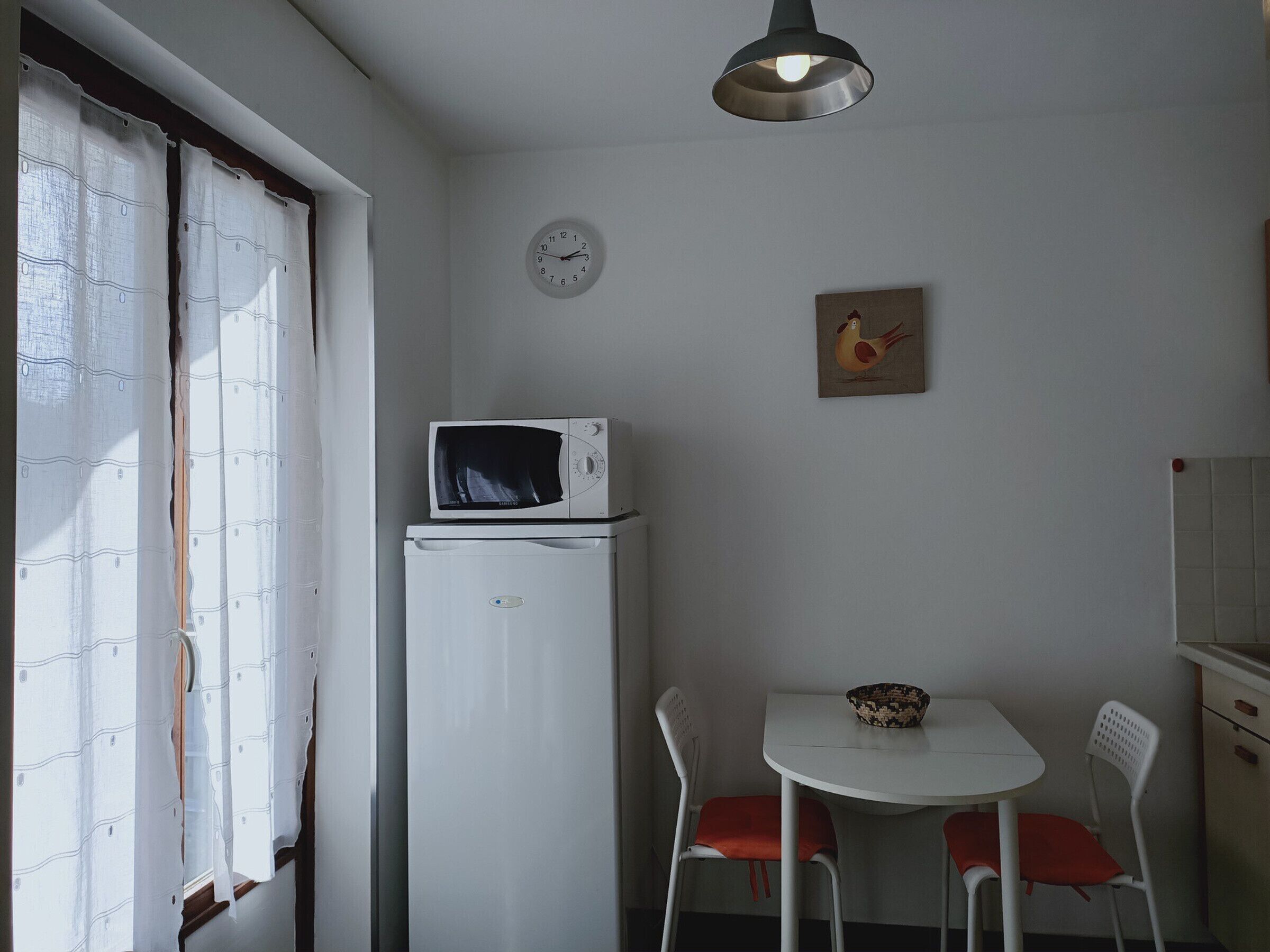 Una piccola cucina con un forno a microonde sopra un frigorifero, un tavolo da pranzo con due sedie, una sveglia a muro e una finestra con tende.