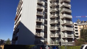 Un edificio residencial de varios pisos con balcones, cielo despejado y coches estacionados al frente.