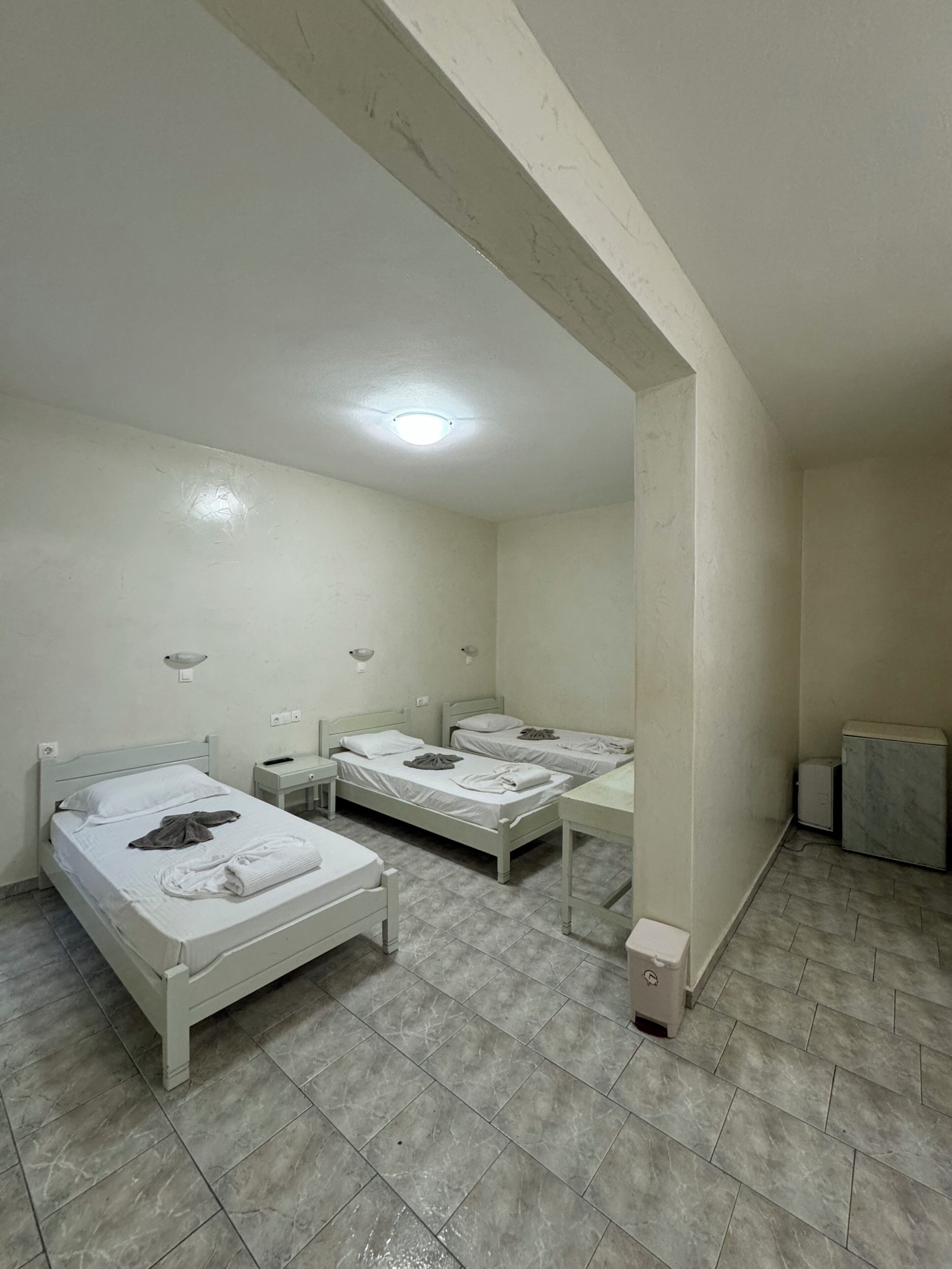 Una habitación con tres camas, una mesita, una lámpara de pared y una luz en el techo.