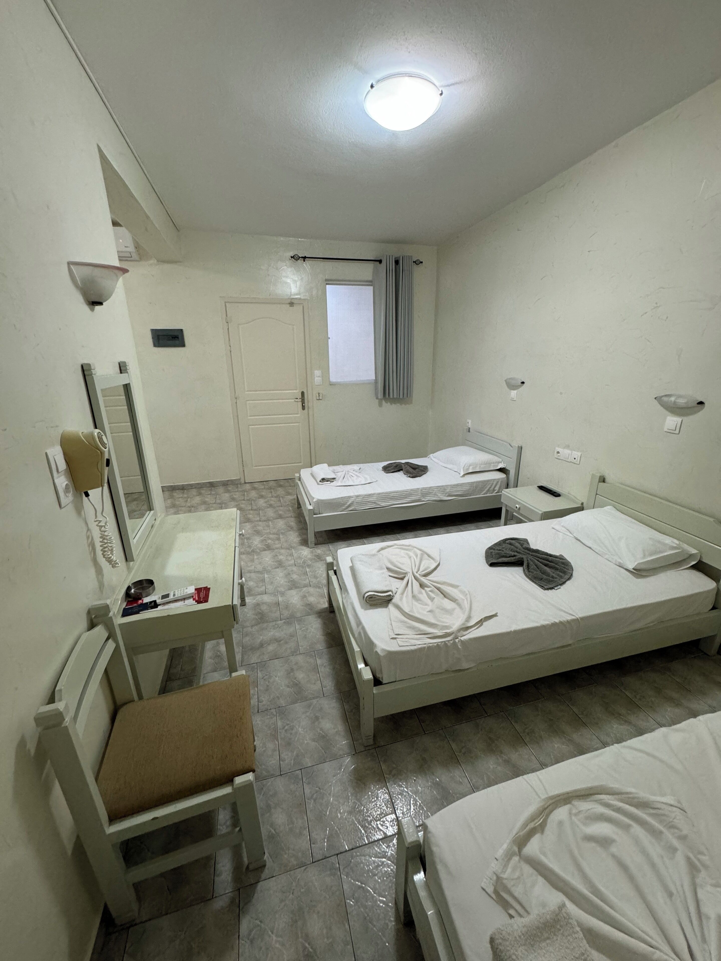 A hotel room with two beds, a small table with a chair, a mirror, and a window with curtains.
