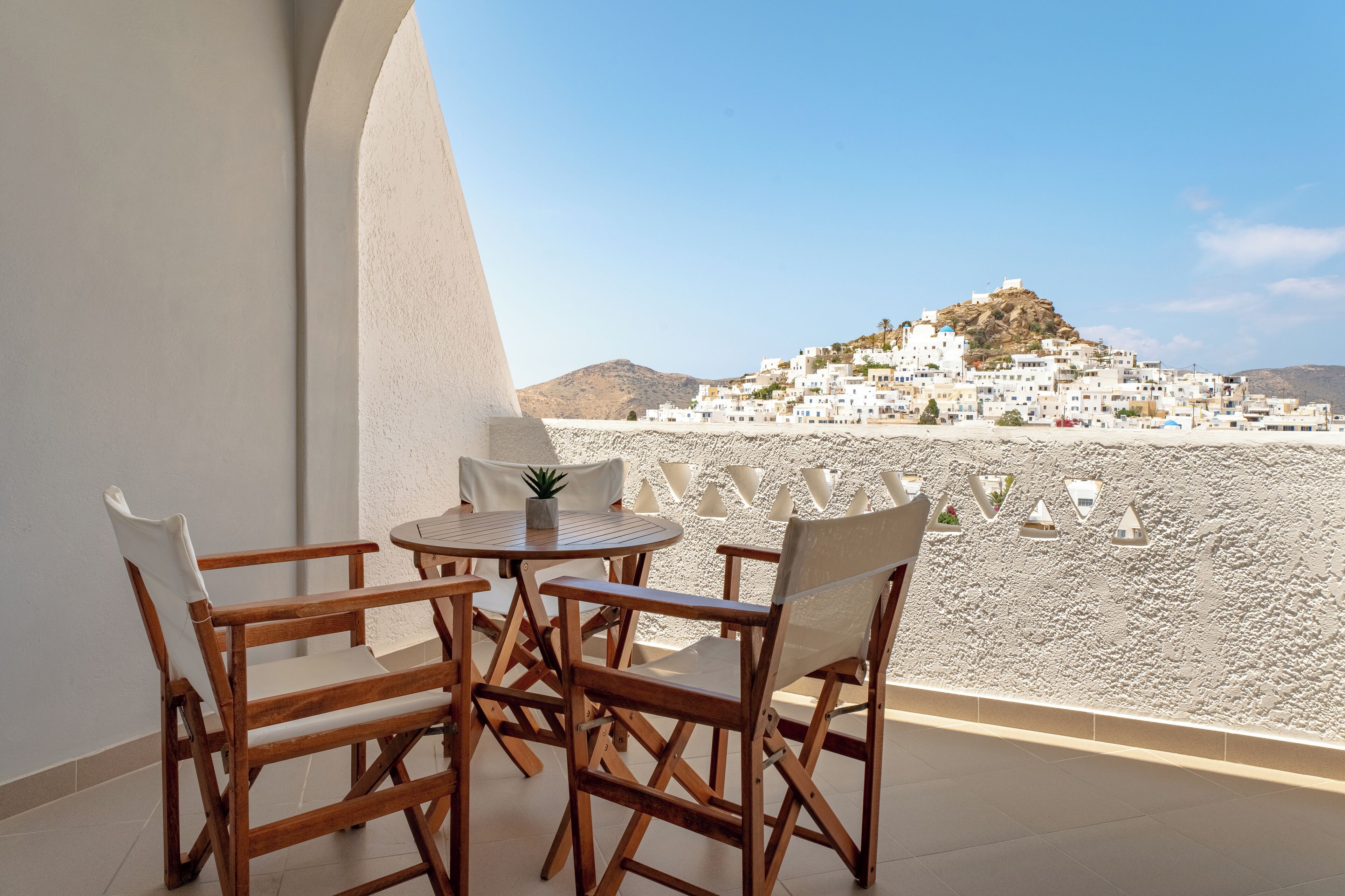 A balcony with a wooden table and chairs, overlooking a white-washed village with a hill in the background.