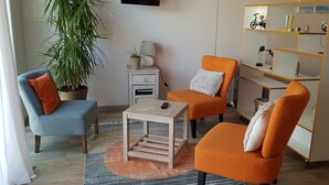 Un salon avec un fauteuil bleu, un fauteuil orange, une petite table, une bibliothèque et une plante en pot.