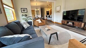 Een moderne woonkamer met een bank, salontafel en televisie. In de achtergrond is een eethoek met stoelen en een kroonluchter.