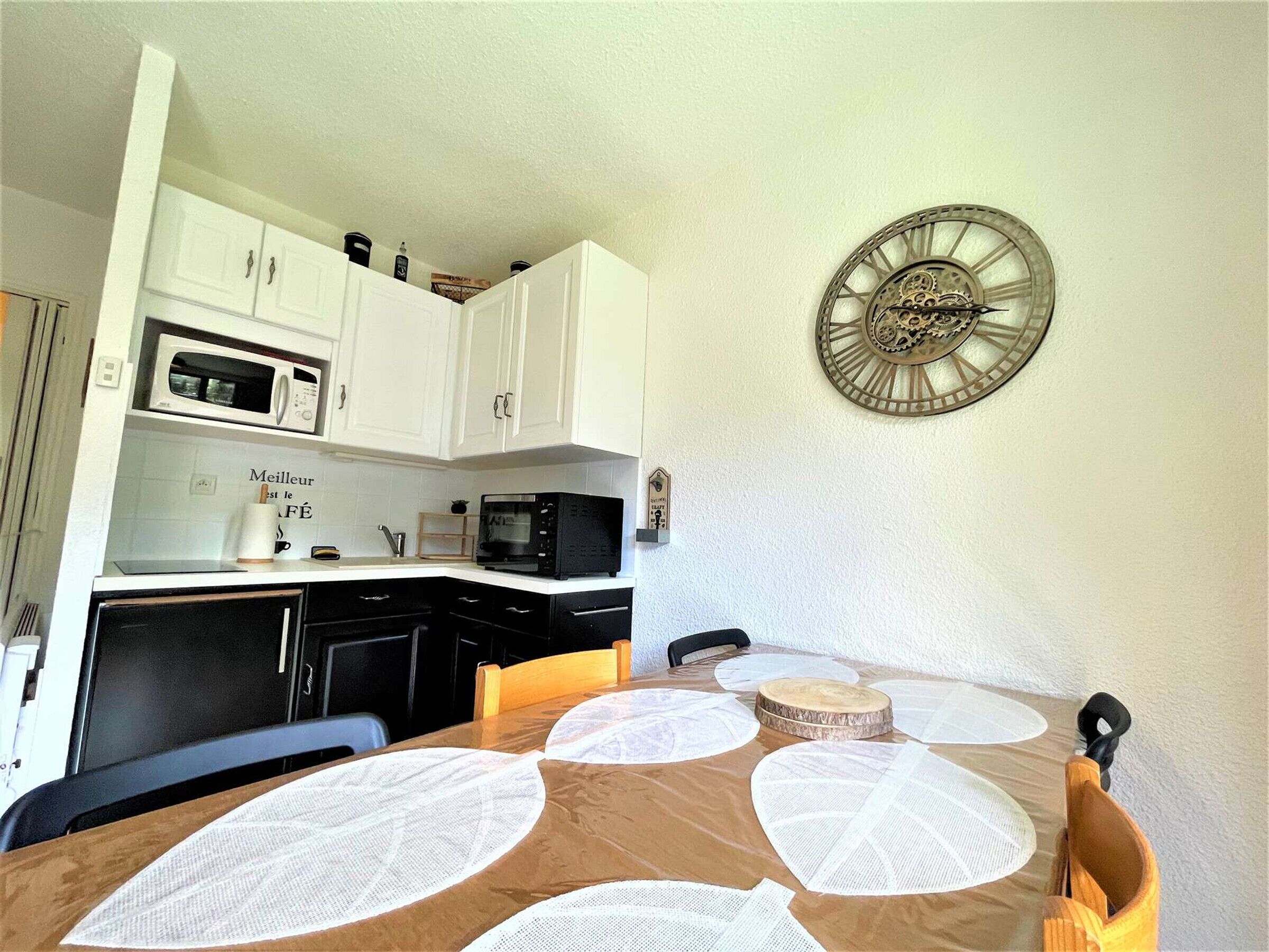 Une cuisine de taille réduite avec des meubles blancs, un four à micro-ondes, une horloge et une table à manger avec des sets de table.