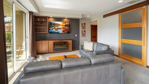 Una sala moderna con un sofá de cuero gris, un televisor de pantalla plana montado en la pared, un mueble de entretenimiento de madera y un ventanal corredero que da a un área exterior.
