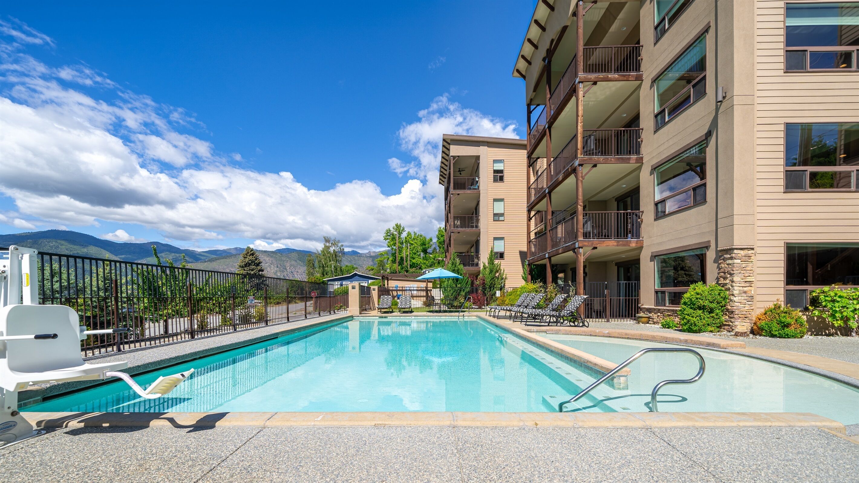 Une piscine avec des chaises longues, un espace délimité par une clôture et un bâtiment doté de balcons.