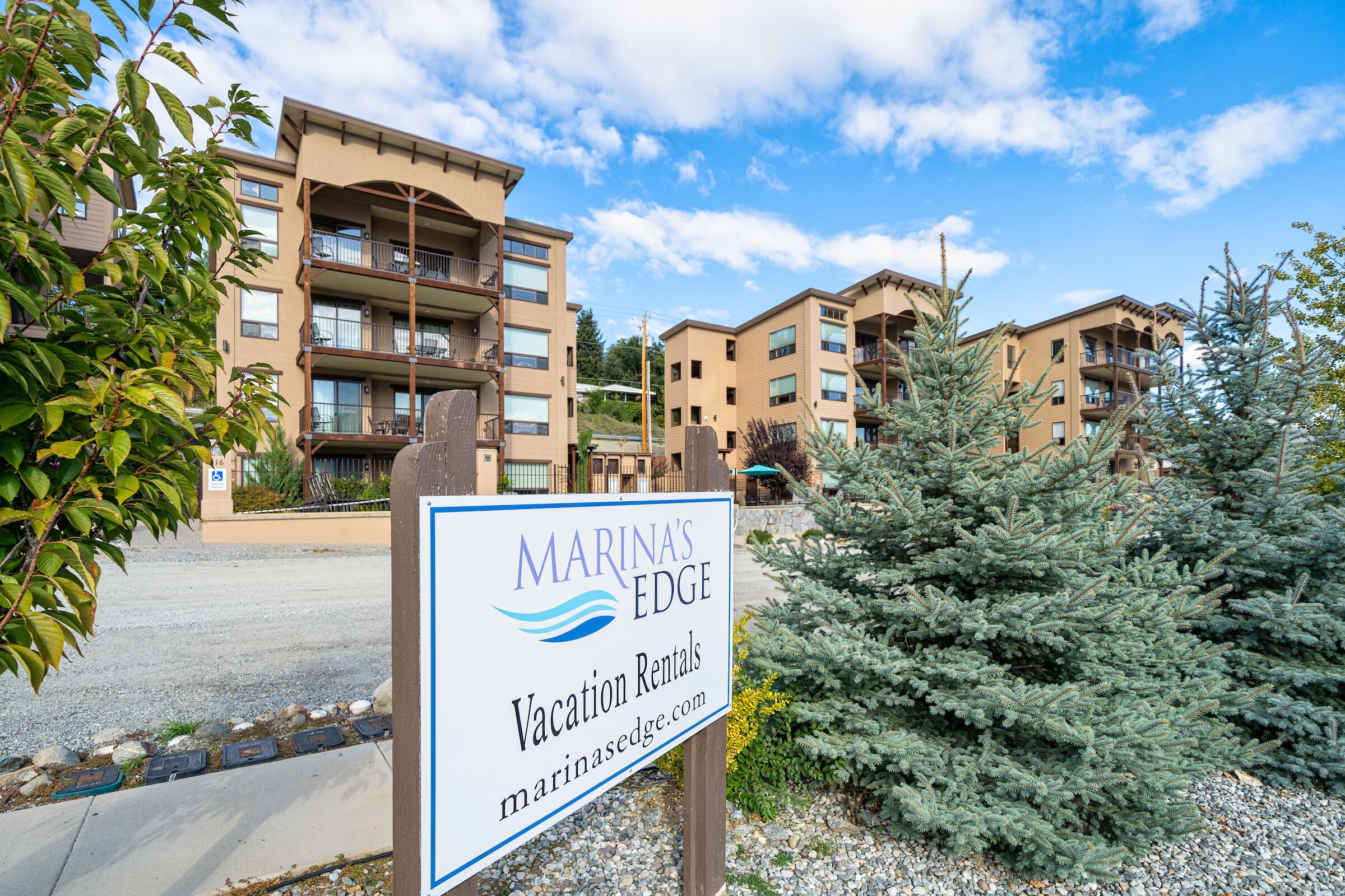A sign for Marina's Edge Vacation Rentals with a view of multi-story buildings and a pine tree in the foreground.