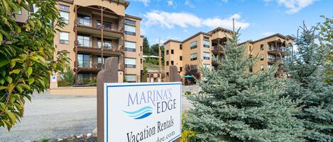 A sign for Marina's Edge Vacation Rentals with a view of multi-story buildings and a pine tree in the foreground.