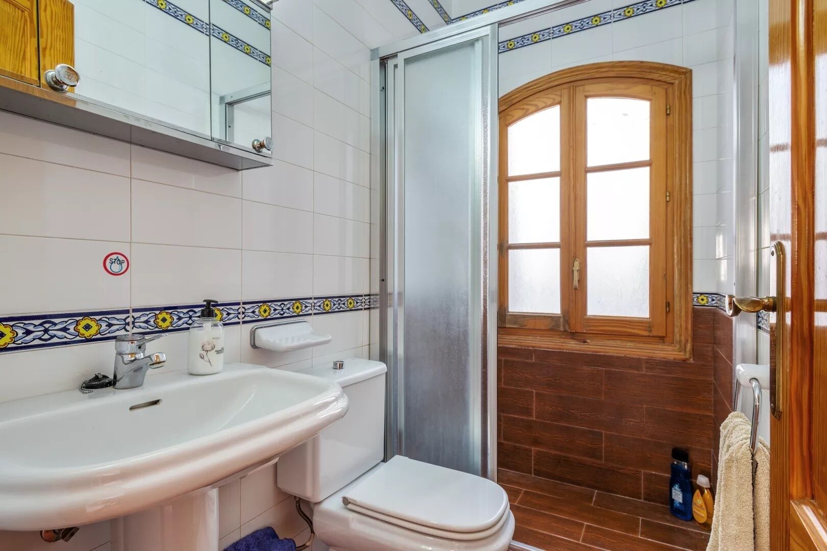 Un baño con lavamanos, inodoro y ducha. Se ve una ventana con marco de madera y una puerta.