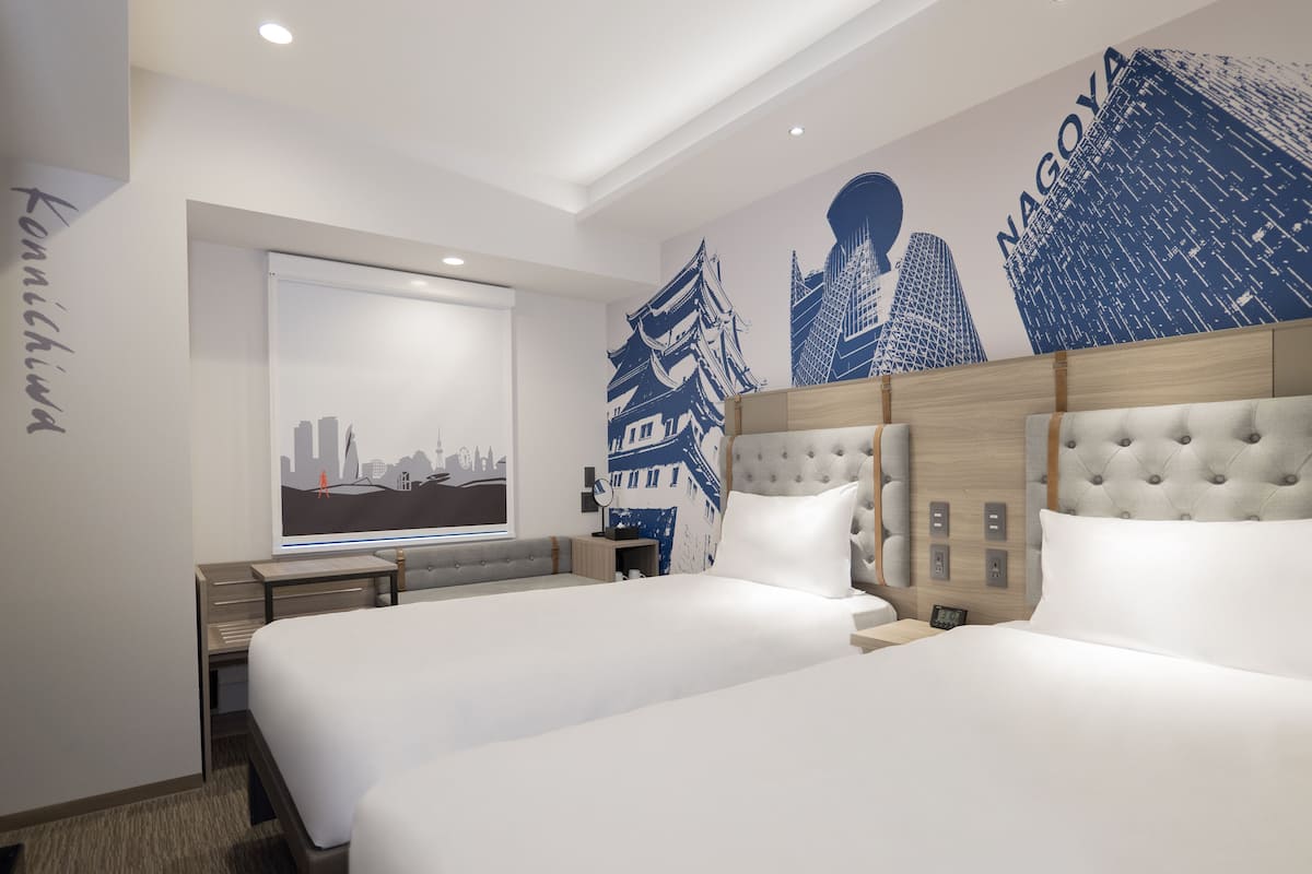 Une chambre d’hôtel avec deux lits, une grande fenêtre donnant sur le paysage urbain et un mur orné d’une fresque représentant la silhouette de la ville.