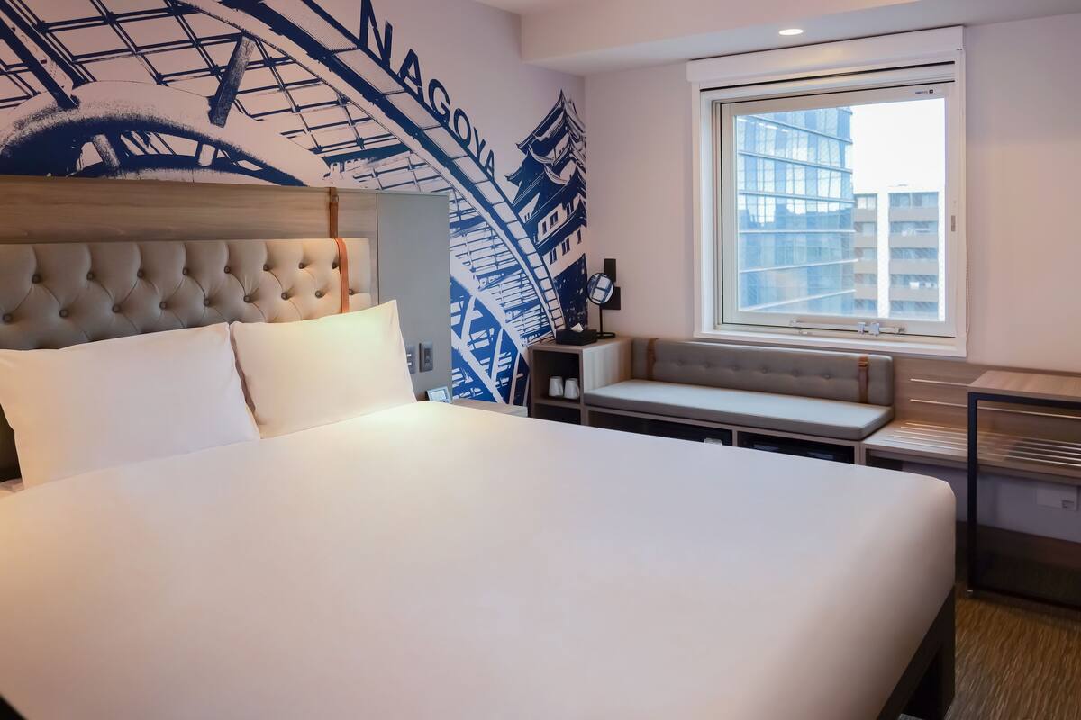 Une chambre d’hôtel avec un grand lit, une tête de lit, une fenêtre donnant sur des immeubles, un mur orné d’un motif bleu et blanc, et une petite table avec une lampe.