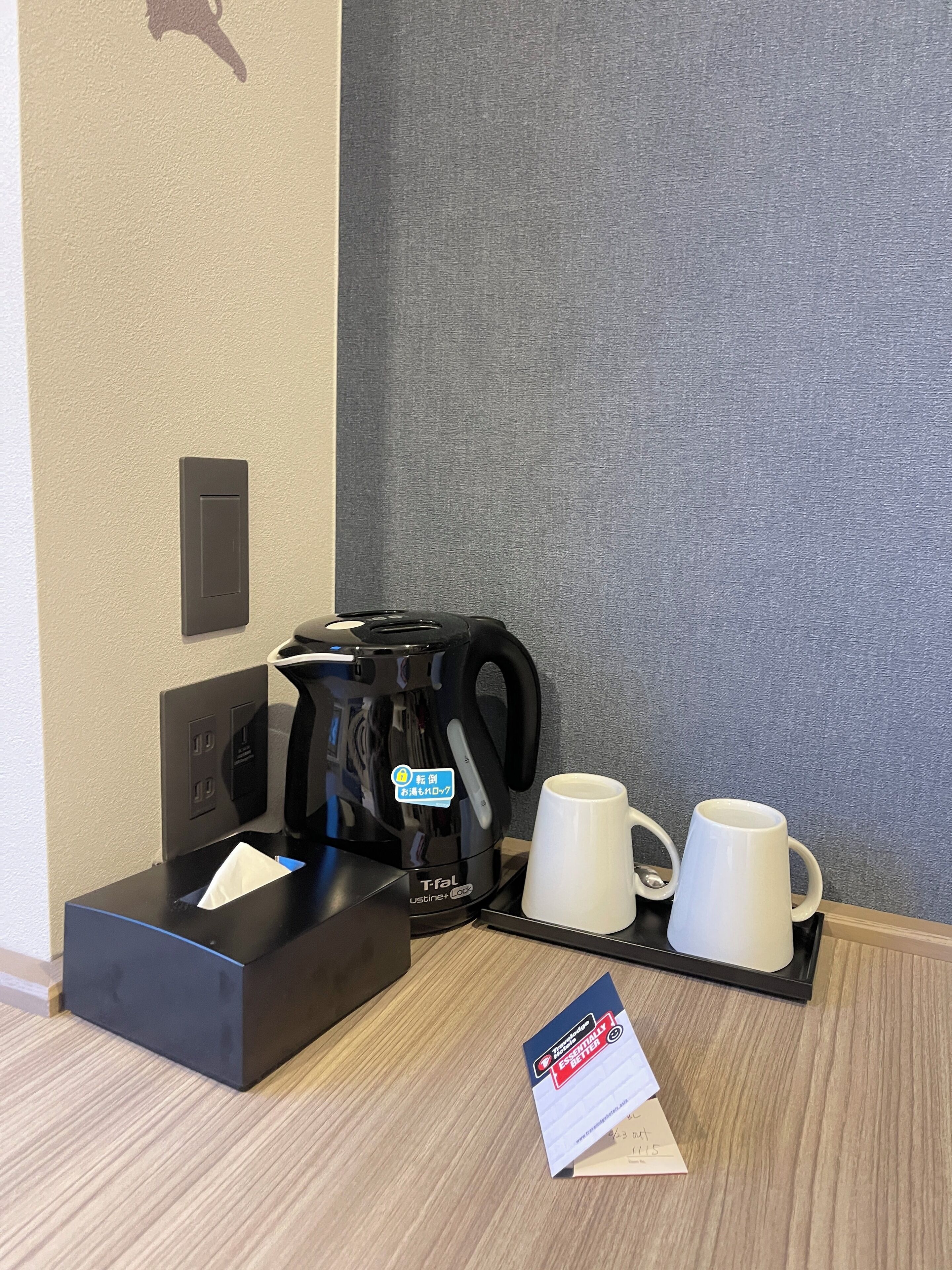 A coffee station with a Tefal coffee maker, two white mugs, a tissue box, and a promotional card on a wooden table.