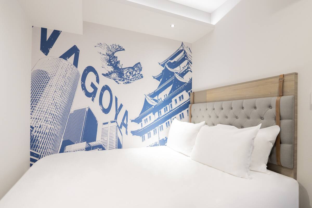 Une chambre d’hôtel moderne dotée d’un grand lit, d’une tête de lit capitonnée et d’une fresque murale représentant un paysage urbain et des bâtiments d’architecture traditionnelle.