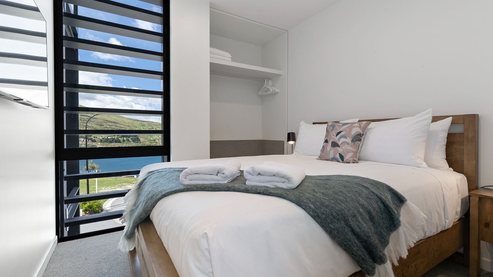 Ein Schlafzimmer mit einem Bett, einem Nachttisch, einem Fenster mit Jalousien und Blick ins Freie.