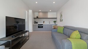 Un salon moderne avec un téléviseur à écran plat, un canapé gris et une cuisine en arrière-plan.