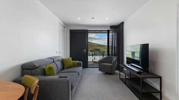 Un salon moderne avec un canapé gris, une petite table, un téléviseur et un balcon offrant une vue sur les collines.