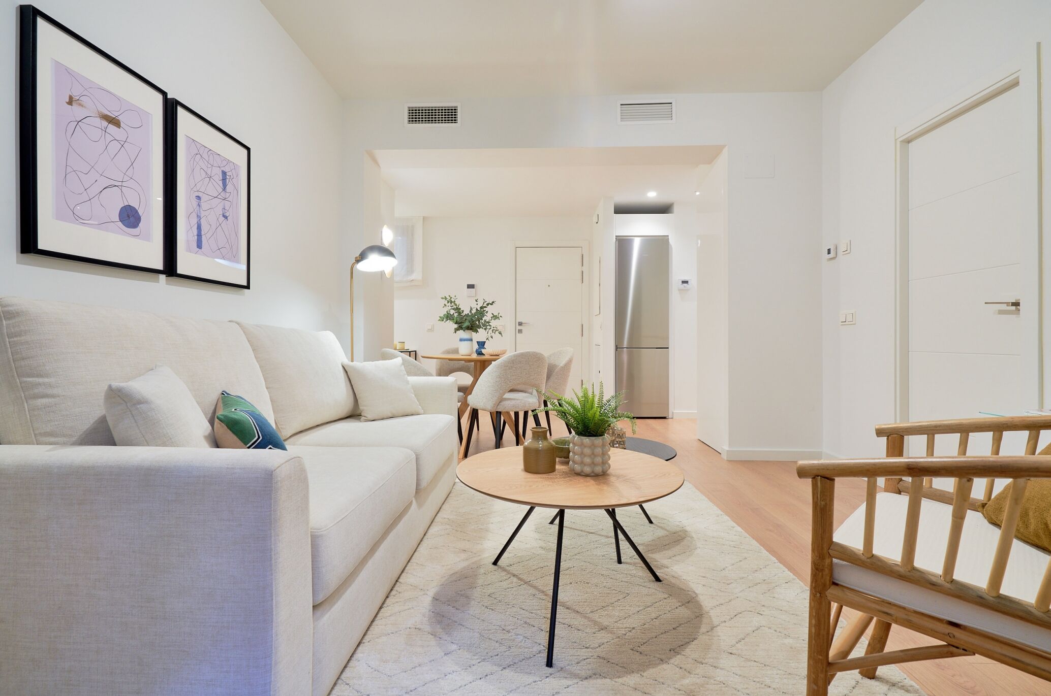 A modern living room with a sofa, a round coffee table, and a wooden chair. There is a door leading to a kitchen area with a refrigerator.