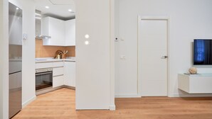 Cozinha moderna com armários brancos, bancada de madeira e forno embutido. Há uma porta que dá para outro cômodo e uma mesa pequena com um objeto decorativo.