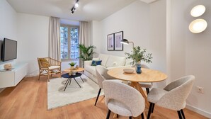 Uma sala de estar moderna com uma mesa de madeira redonda, cadeiras estofadas em cinza, um sofá branco e uma TV sobre um móvel branco.