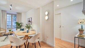 Uma sala de estar moderna com uma mesa redonda de madeira, duas poltronas bege, um vaso com planta e um sofá.