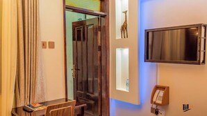 A hotel room with a bed, a desk with a chair, a telephone, and a television mounted on the wall. - Imara Hotel (Kampala)