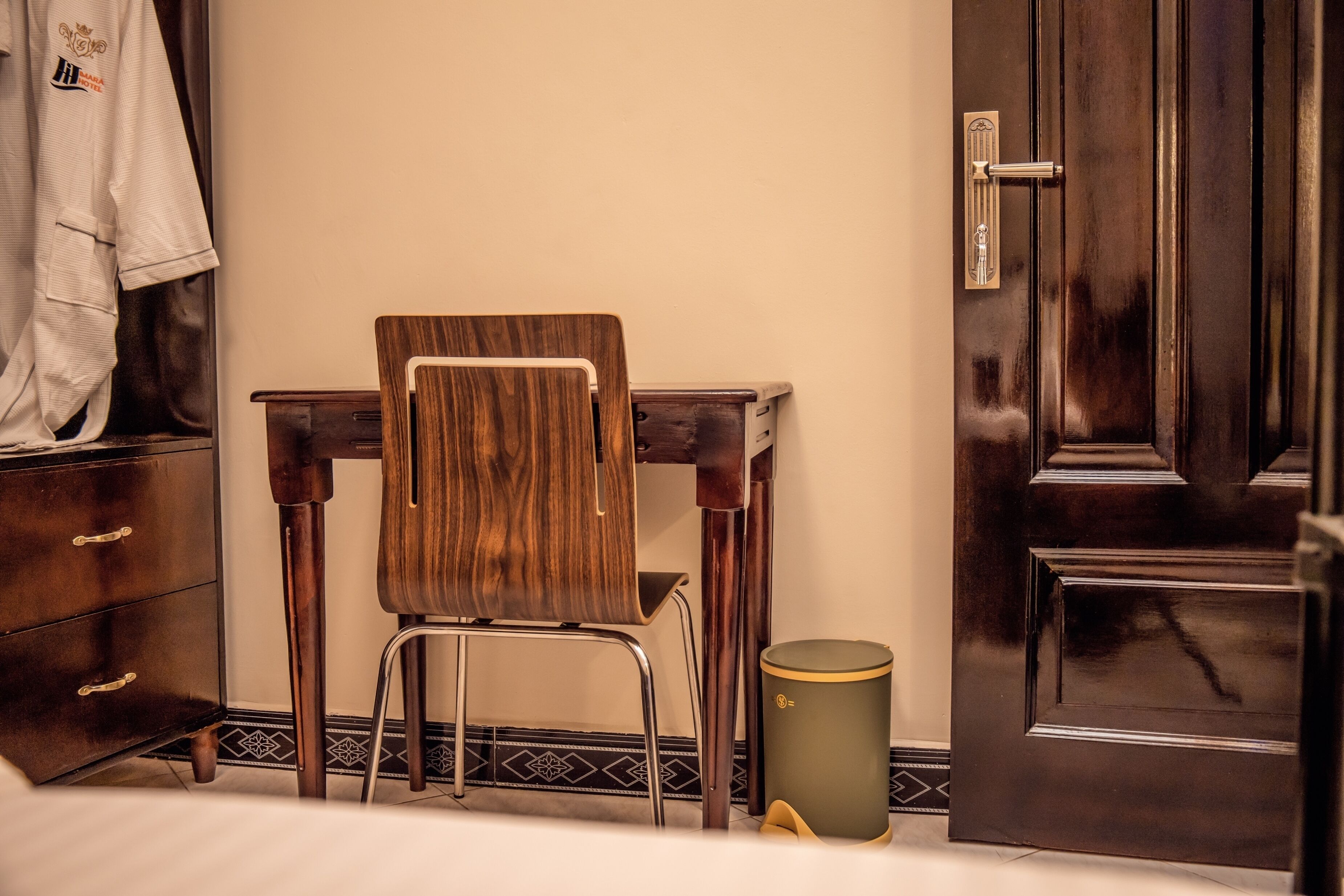 A wooden desk chair with a metal frame, a dark wooden dresser with drawers, a closed door, and a trash can.