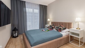 Ein modernes Hotelzimmer mit einem großen Bett, zwei Nachttischen mit Lampen, einem an der Wand befestigten Fernseher und einem Fenster mit grauen Vorhängen.