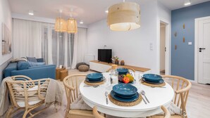 Een moderne woonkamer met een ronde eettafel gedekt voor vier personen, een blauwe bank, rieten stoelen en een televisie op de achtergrond.