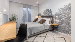 Een moderne slaapkamer met een bed, een bureau en een stoel. Er is een raam met gordijnen, een muur met een boommuurdecoratie, en een kleine tafel met een lamp.