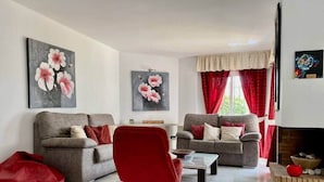 A living room with two grey sofas, a red chair, and a coffee table. There are floral paintings on the walls and a fireplace with a red curtain. - House Pauline 3 Bed. Poolside, Private Garden. M-5 (Marbella)