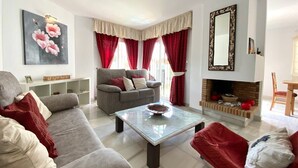 A living room with a sofa, armchair, and a coffee table. There is a fireplace with red curtains and a painting on the wall. - House Pauline 3 Bed. Poolside, Private Garden. M-5 (Marbella)