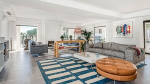 A modern living room with a sofa, armchair, coffee table, and ottoman. There is a fireplace, a dining area, and a large window with a view. - Contemporary Golf Villa Las Brisas Nueva Andalucia (Marbella)