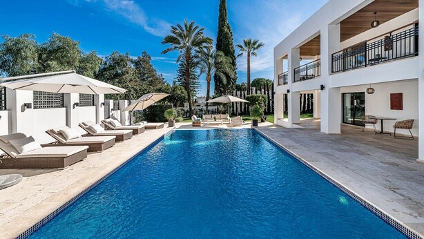 A swimming pool with lounge chairs, a covered seating area, and a two-story building with balconies. - Contemporary Golf Villa Las Brisas Nueva Andalucia (Marbella)