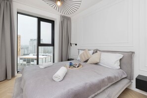 Ett modernt hotellrum med en stor säng, utsikt över staden genom stora fönster som sträcker sig från golv till tak och en minimalistisk design.
