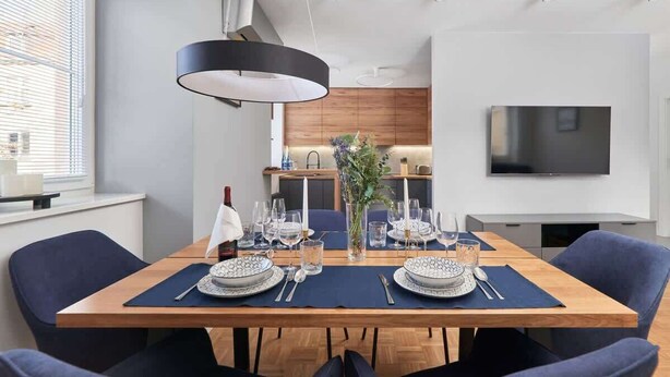 Ein moderner Essbereich mit einem Holztisch, gedeckt für vier Personen, umgeben von dunkelblauen Stühlen. Eine große schwarze Pendelleuchte hängt darüber, und ein Flachbildfernseher ist rechts an der Wand montiert.