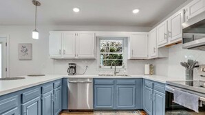A modern kitchen with white and blue cabinets, a stainless steel dishwasher, and a window providing natural light. - Restful Haven Near Savannah - King Bed (Pooler)